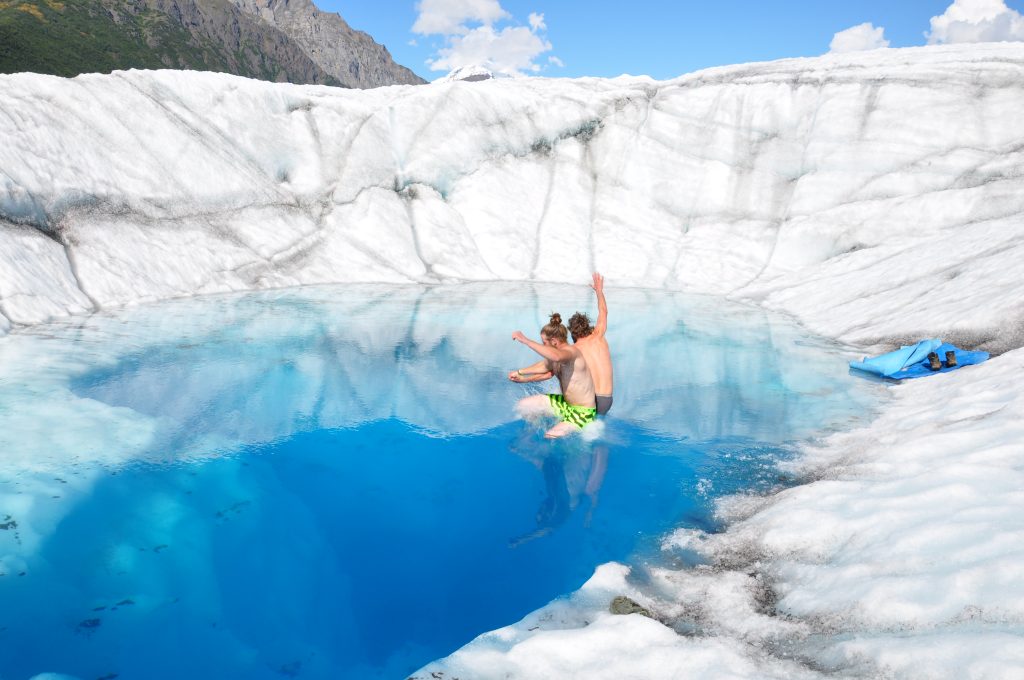 Two people holding hands and jumping into a blue pool on the Root Glacier