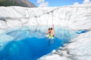 Two people holding hands and jumping into a blue pool on the Root Glacier