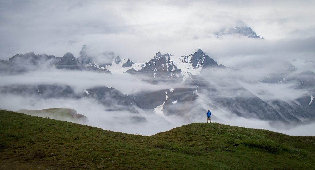 A person standing on rolling green hills admiring the clouds rolling over the Wrangell Mountains