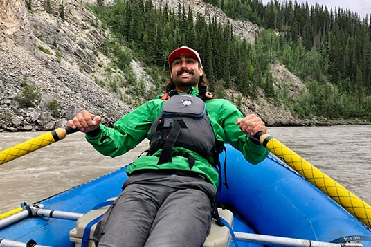 A smiling rafting guide pulls back on the oars of a blue raft on a glacial river in Alaska
