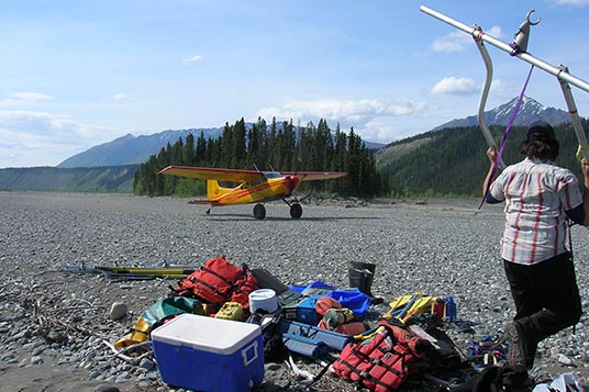 A yellow bush plane lands on a gravel river bar next to a pile of rafting gear in Wrangell - St. Elias National Park