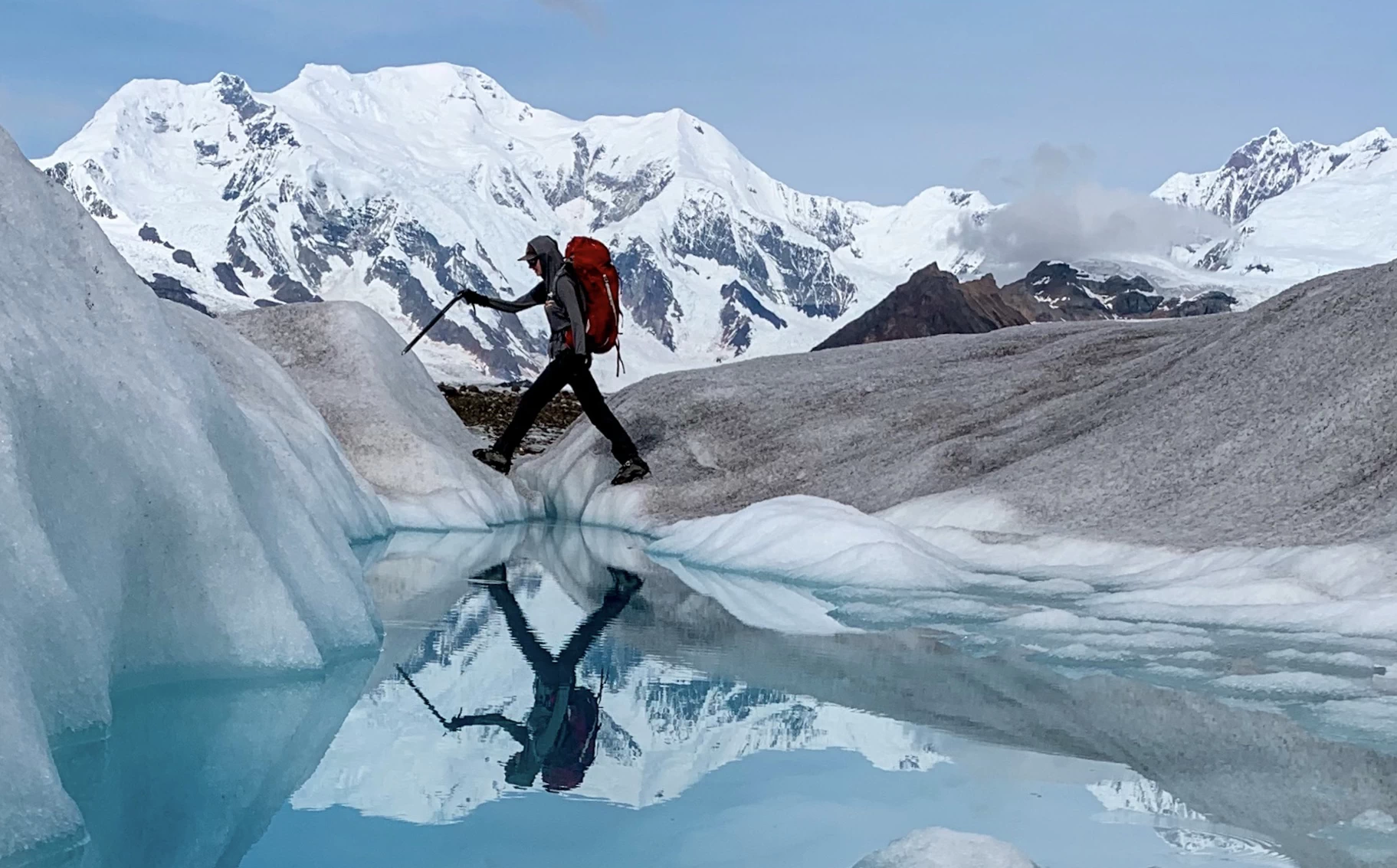 Glacier hiker steps over reflective pool with Mt. Blackburn in the background