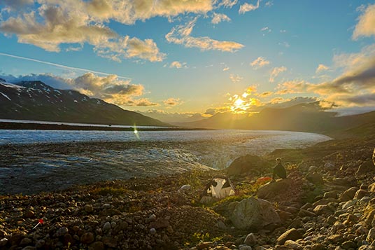 A backpacker and her tent on rocks at sunset at a glacier's edge