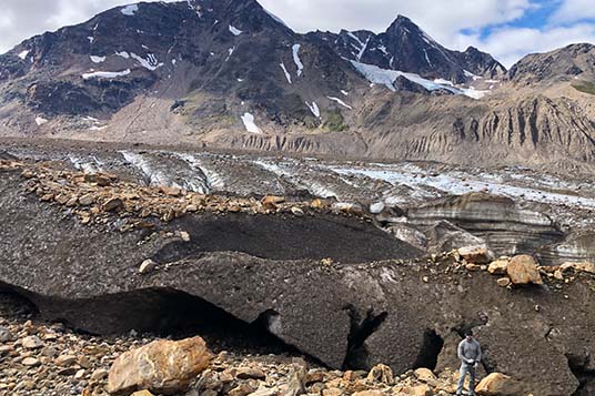 A hiker stands on rocks in front of a massive and dirt-covered glacier