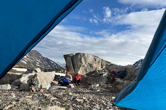 A view out of a tent to hikers resting against rocks