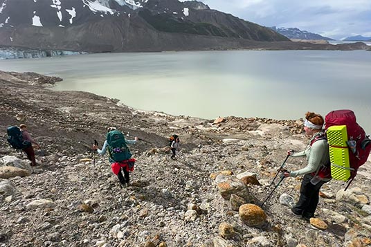 Backpackers move downhill through rocky terrain to a lake