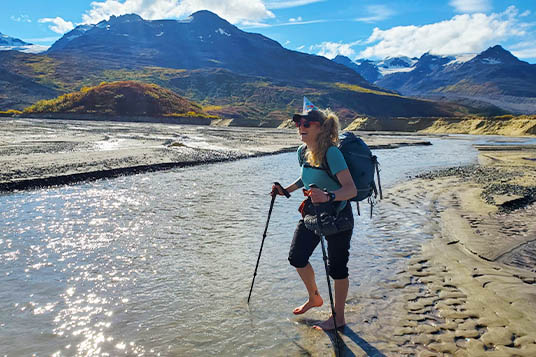 A hiker in a party hat digs her toes into the sand of a shallow stream in the mountains