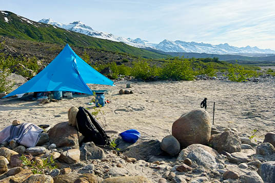 A tent is set up on sand with snow-covered mountains in the background