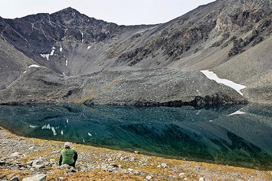 A woman sits on the tundra facing a giant, dark, clear alpine lake