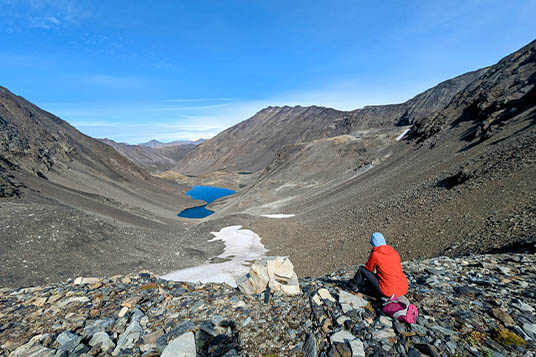 A hiker sits on a high rocky pass overlooking a bright blue lake