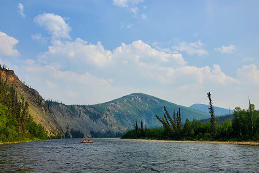 A calm scene of a river with a small raft in front of a mountains in backcountry Alaska
