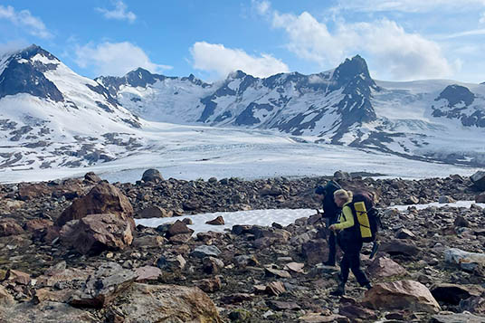 Two backpackers walk on rocks in front a glaciated peak