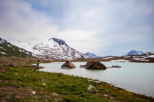 A lone backpacker stands at the edge of an alpine lake in the Chugach Mountains