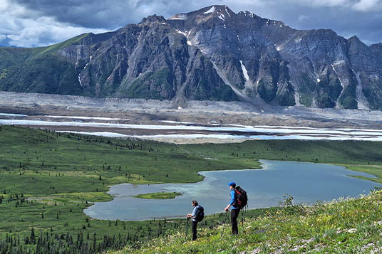 Two people on a slope overlooking a blue lake and the Kennicott Glacier