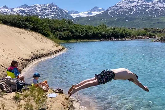 A man jumps into a lake in the mountains while kids wait on the sandy beach