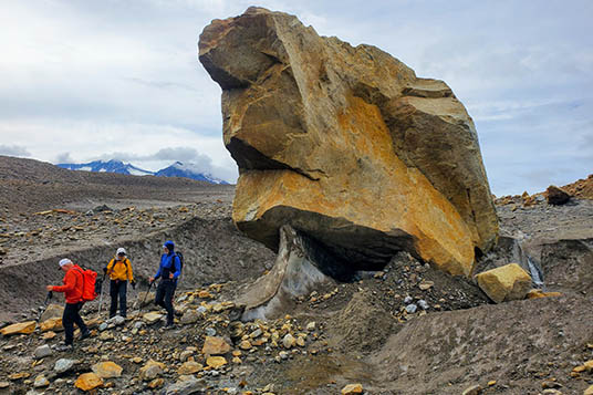 Hikers in bright jackets walk on a glacier next to an enormous boulder