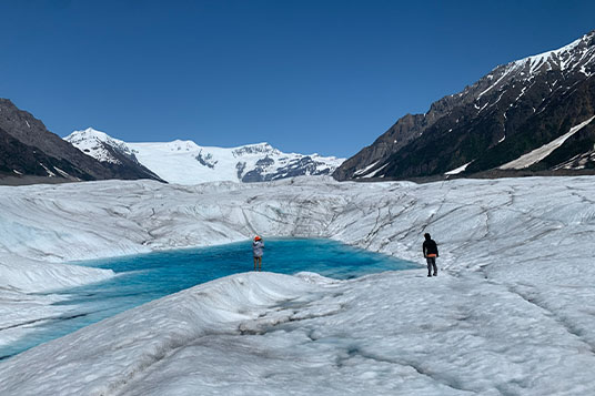 Two hikers approach a large blue pool on the Root Glacier with the Stairway Icefall behind them