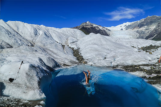 A pair of feet stick out of a blue pool on a glacier that someone has just jumped into