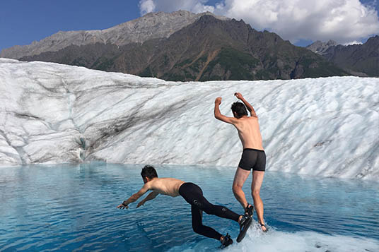 Two men dive into a clear blue pool on a glacier