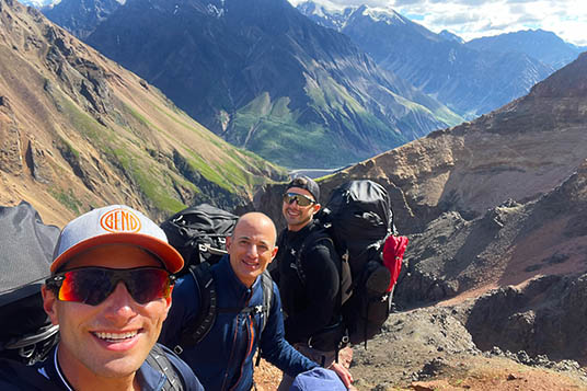 Three backpackers smile against mountainous terrain