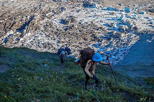 Two backpackers move up a steep slope in front of a field of stranded icebergs
