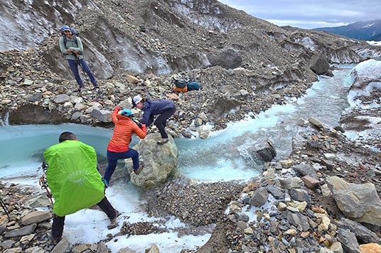 Hikers cross a glacial river on a large boulder