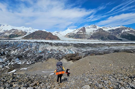Backpackers descend a rocky slope to a cracked up glacier