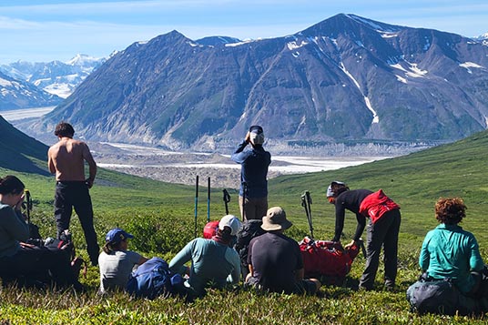 A group of hikers eat lunch on the grass of a high mountain pass
