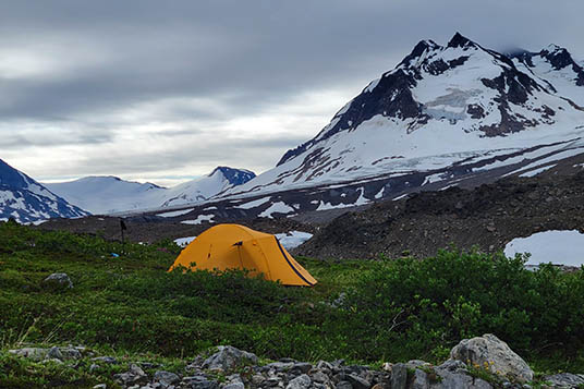 A yellow tent set up on tundra in front of snowy mountains