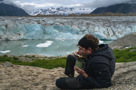 A man eats a backcountry meal with a glacier in the background