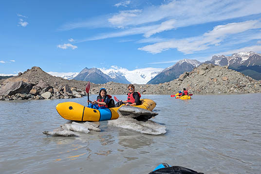 A parent and child paddle a tandem kayak on the Kennicott Glacial Lake beneath the Stairway Icefall
