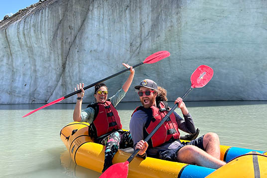 Two smiling paddlers hold their paddles out of the water in a tandem inflatable kayak in front of a huge iceberg