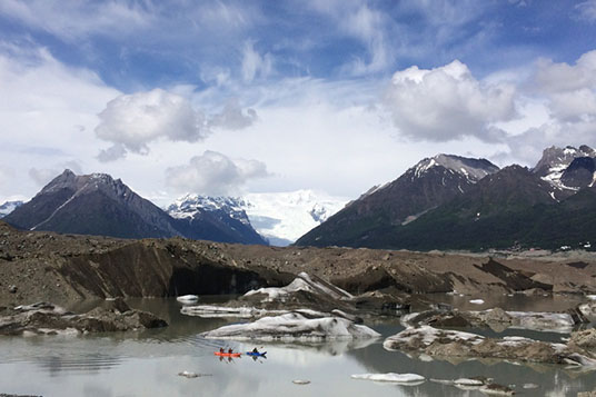 Tiny kayakers in bright boats paddle on the calm waters of the Kennicott Glacier Lake