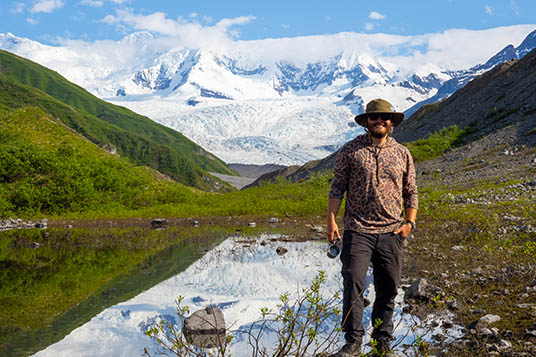 A hiker poses in front of a lake and snowy mountains