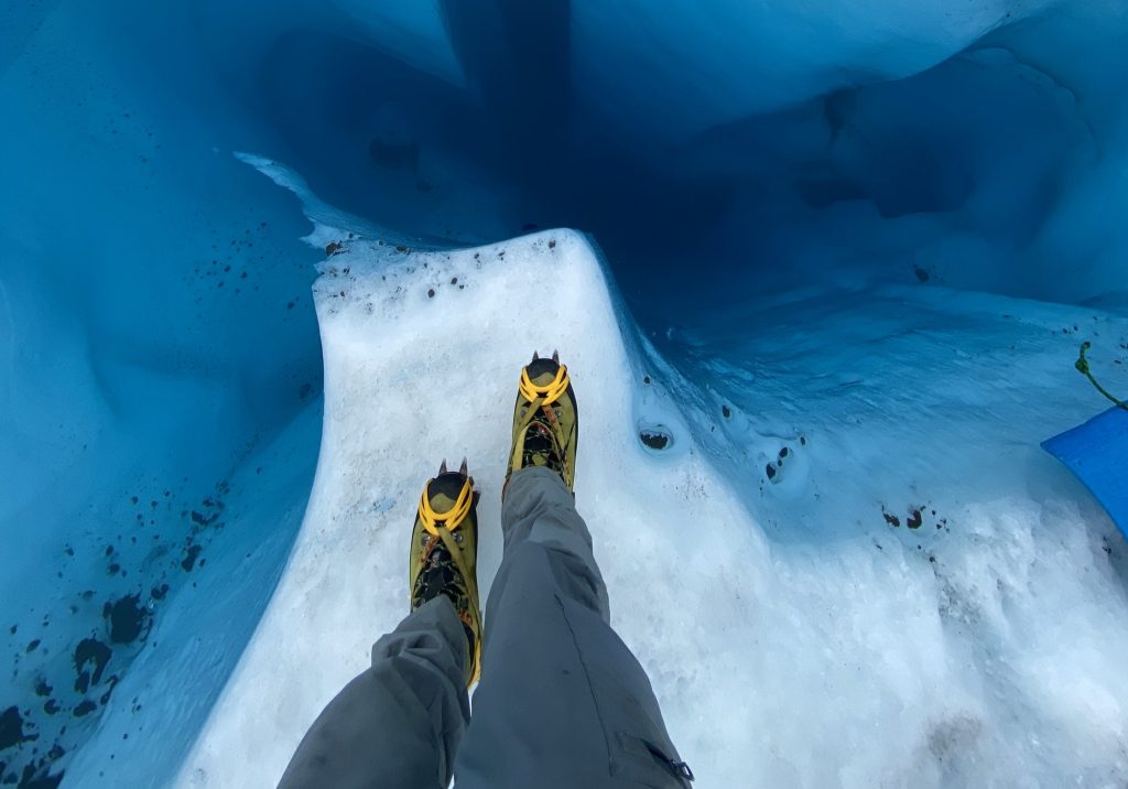 Looking down at legs in crampons at the edge of a glacial pool