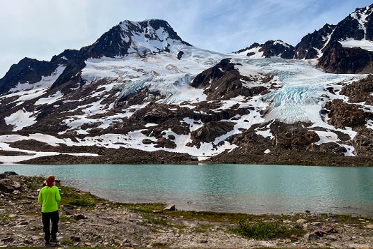 A man stands in front of an alpine lake surrounded by glaciated peaks
