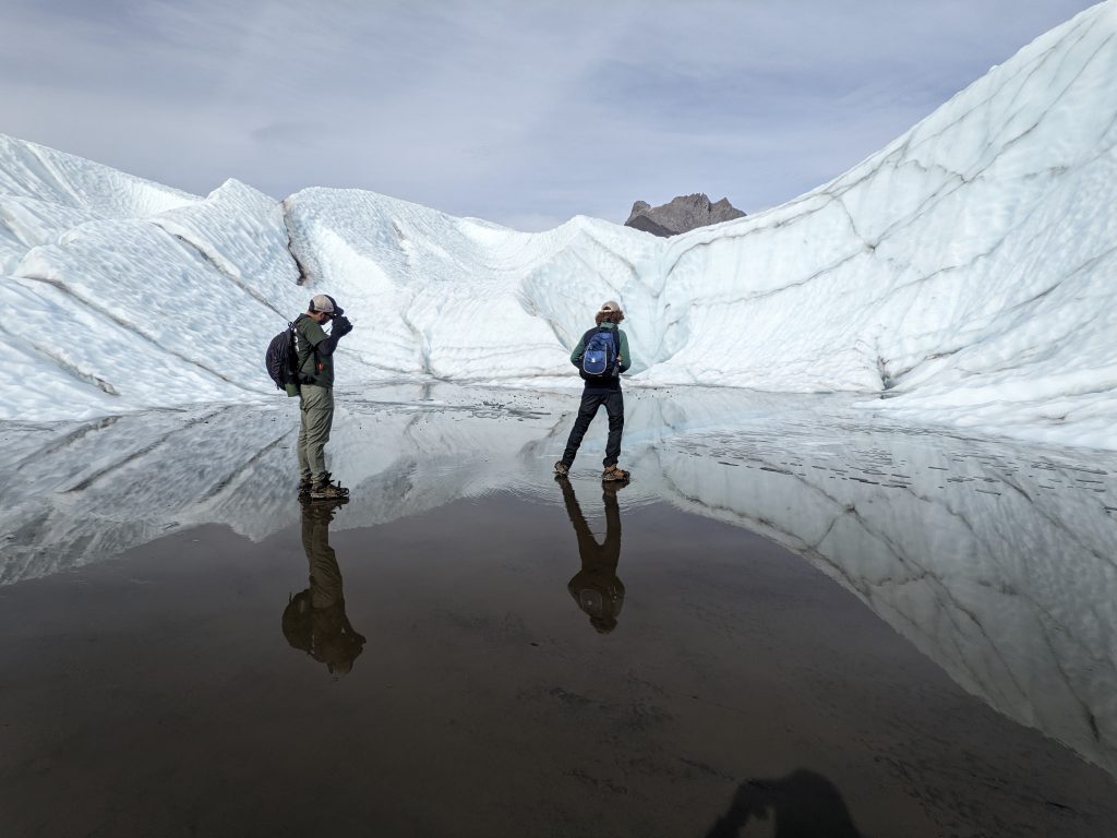 Hikers on a glassy glacial lake in front of a glacier wall