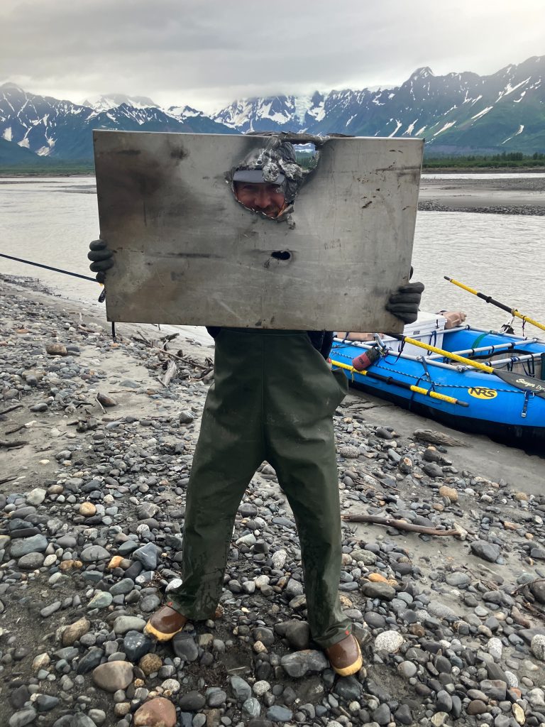 Wilderness guide smiles through lid of tin firepan with blue rafts and snowy mountains in background