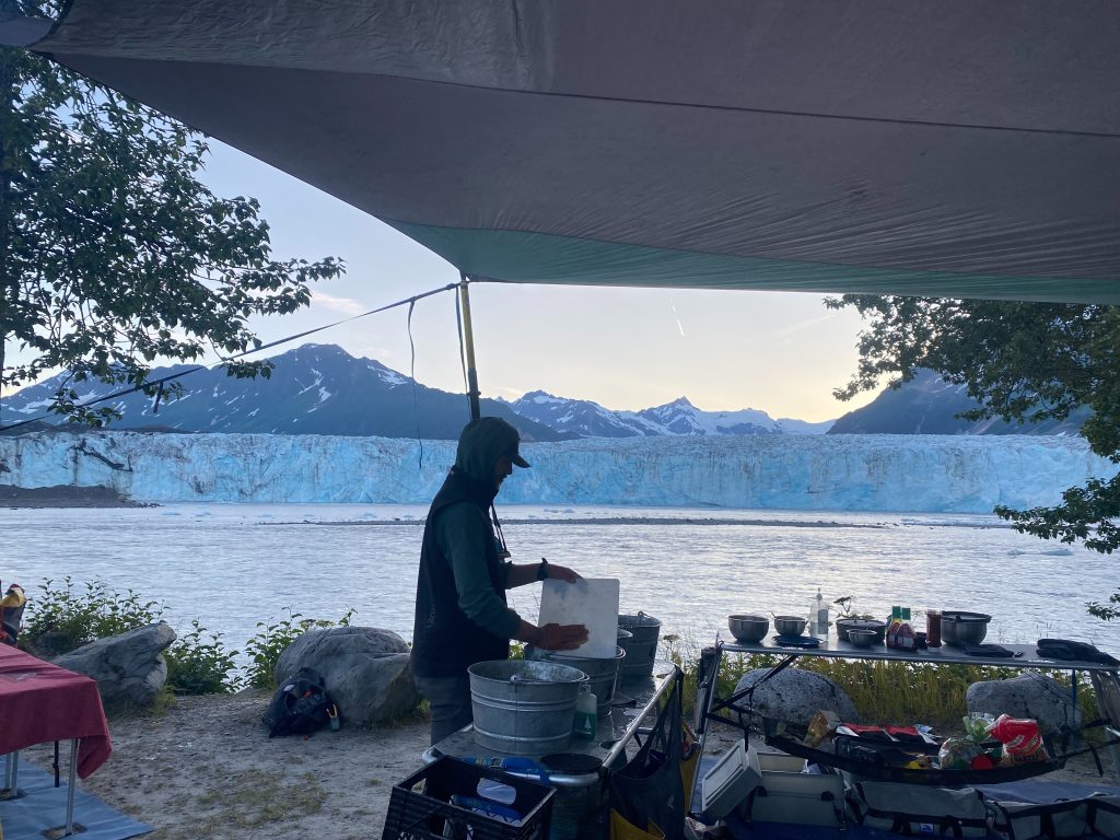 backcountry guide washes dishes in rafting kitchen across from Child's Glacier